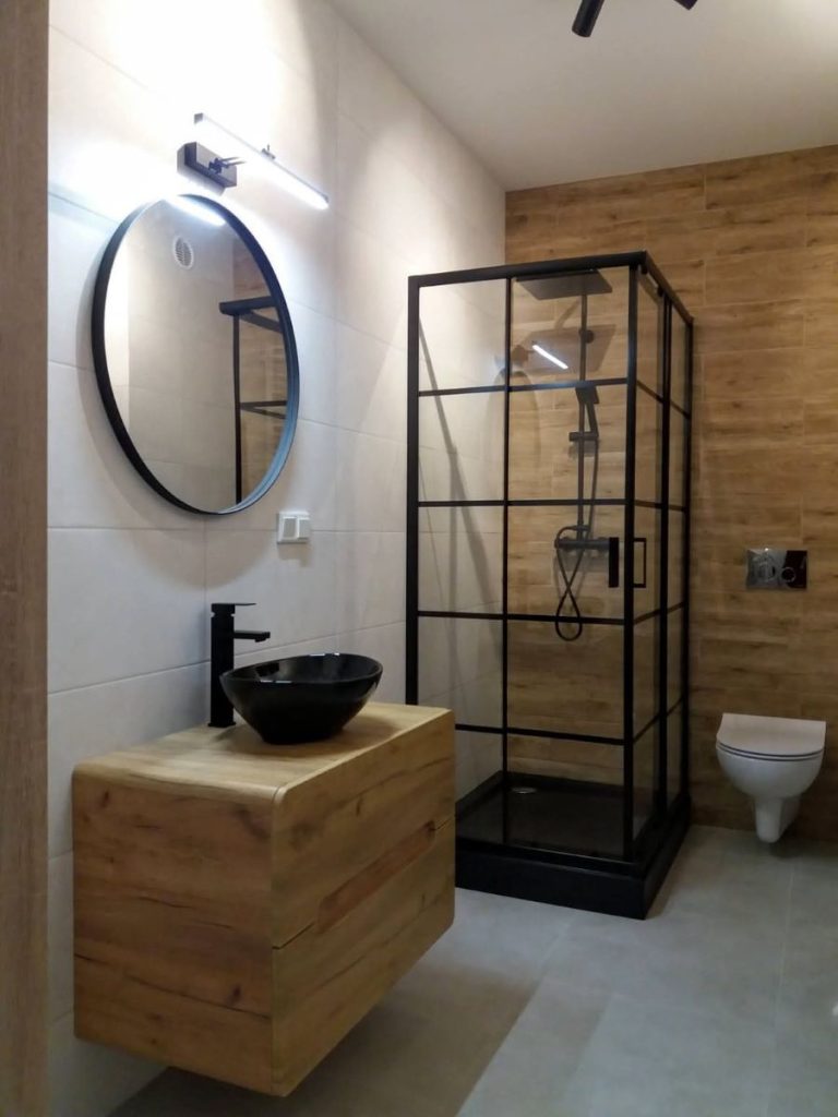 Nowoczesna łazienka w stylu loftowym, czarna kabina prysznicowa typu walk-in, umywalka nablatowa na drewnianej szafce, płytki imitujące drewno na ścianie.