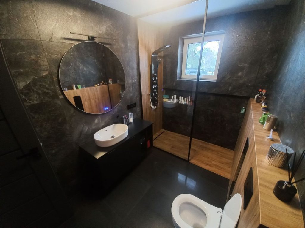 Nowoczesna łazienka w stylu loftowym, czarna kabina prysznicowa typu walk-in, umywalka nablatowa na drewnianej szafce, płytki imitujące drewno na ścianie.