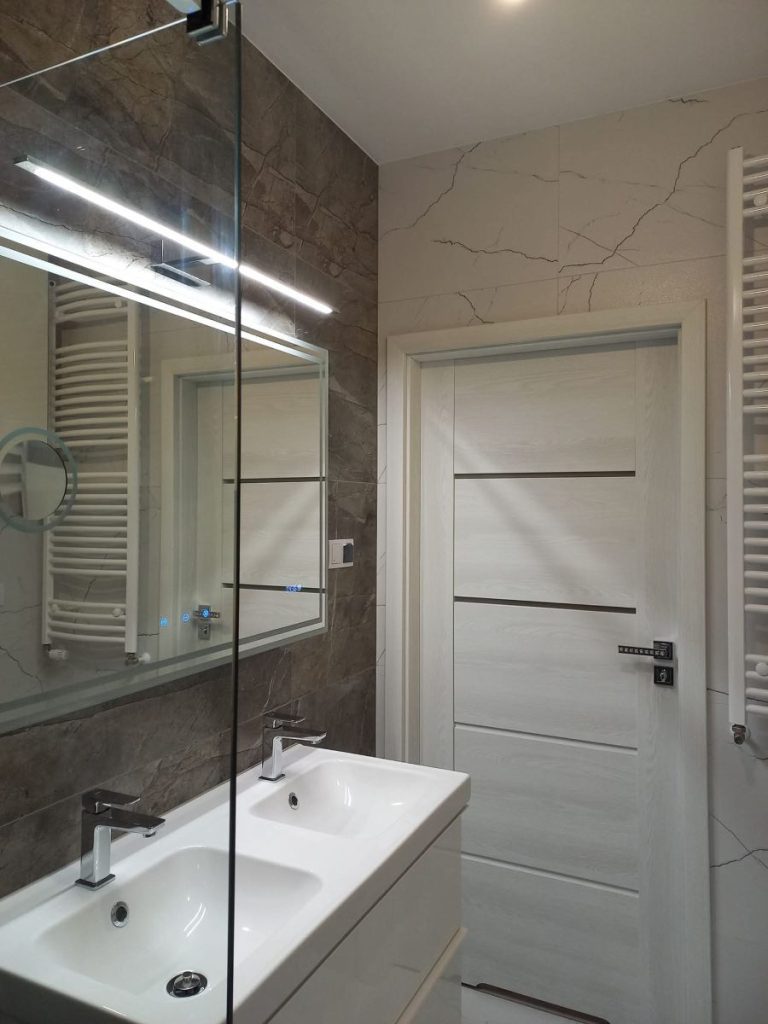 Nowoczesna łazienka, remont łazienki, biała umywalka, duże lustro z oświetleniem LED, beżowe płytki ścienne, minimalistyczny styl.