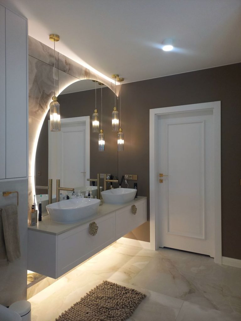 Nowoczesna łazienka w stylu glamour, układanie płytek imitujących marmur, lustro okrągłe LED, złota armatura i dodatki, szafka łazienkowa.