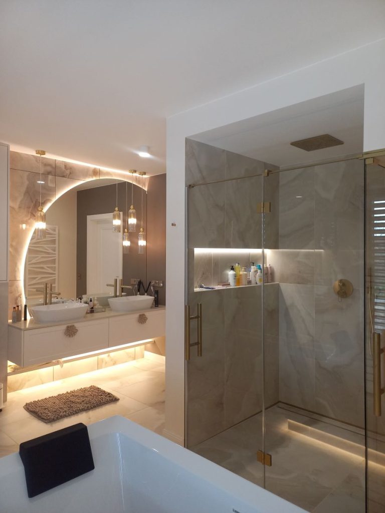 Nowoczesna łazienka w stylu glamour, układanie płytek imitujących marmur, lustro okrągłe LED, złota armatura i dodatki, szafka łazienkowa.