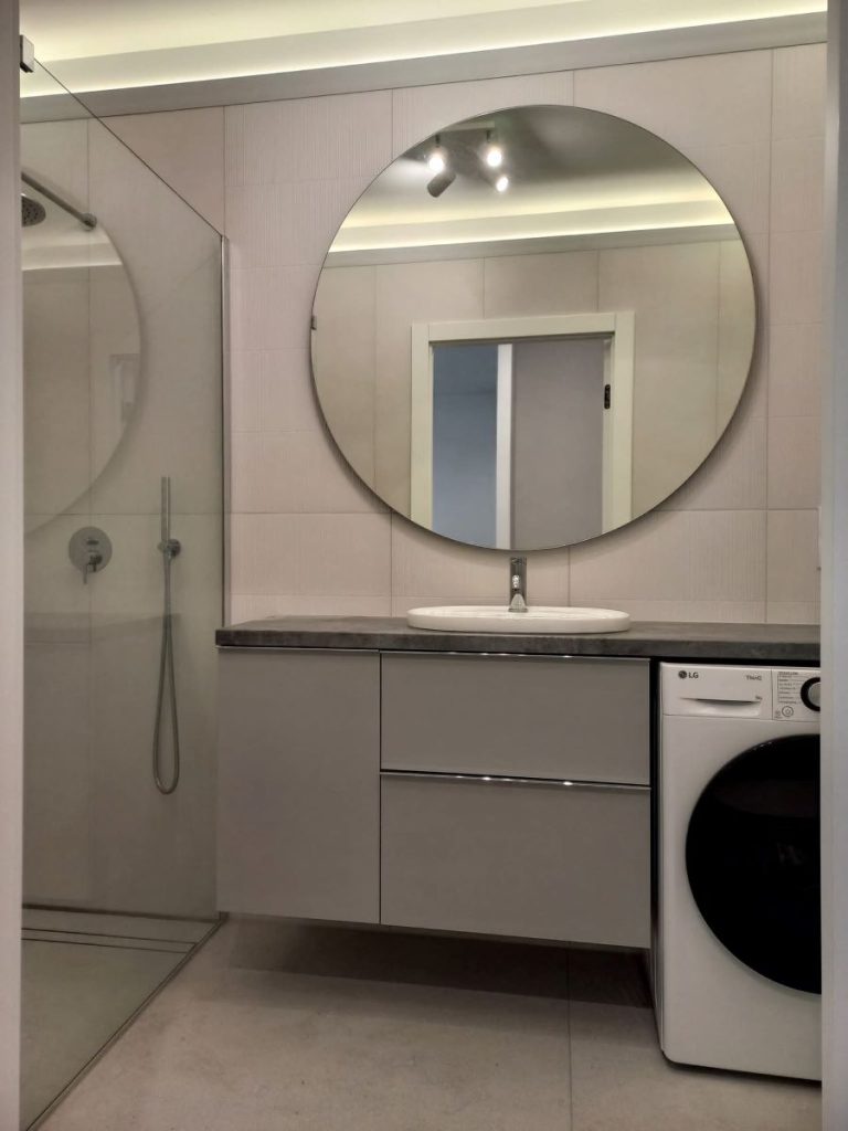 Nowoczesna łazienka w jasnych odcieniach szarości z okrągłym lustrem LED nad szafką umywalkową i pralką wolnostojącą.