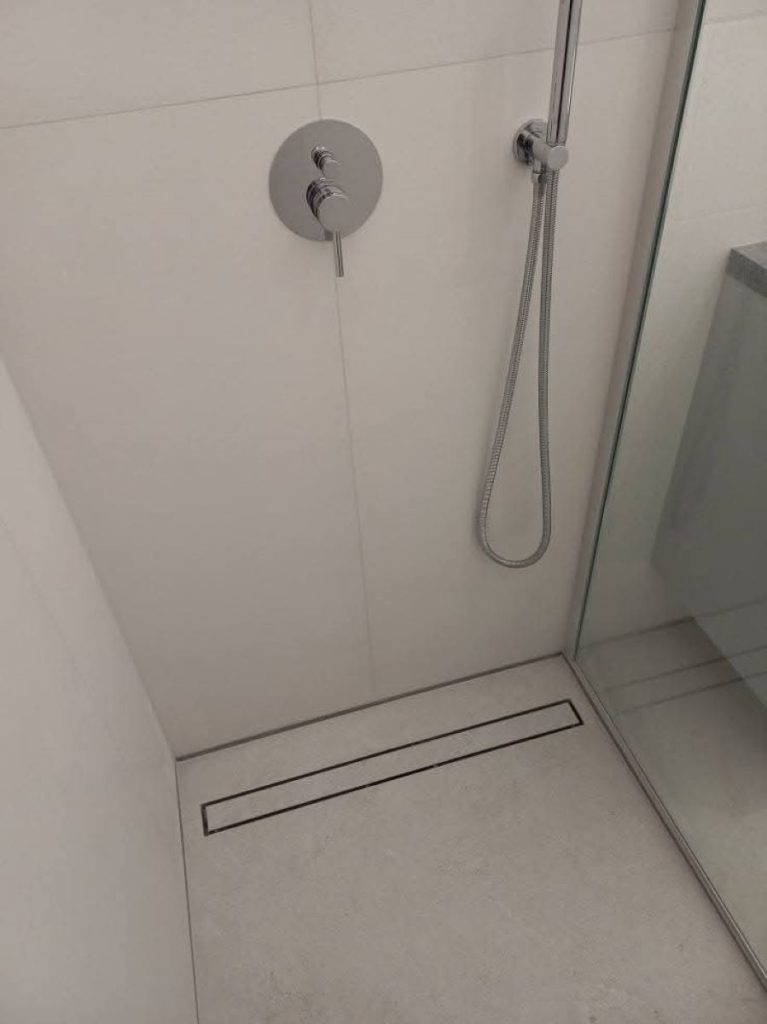 Nowoczesny prysznic walk-in z odpływem liniowym ACO ShowerDrain C w podłodze wyłożonej białą płytką ceramiczną.